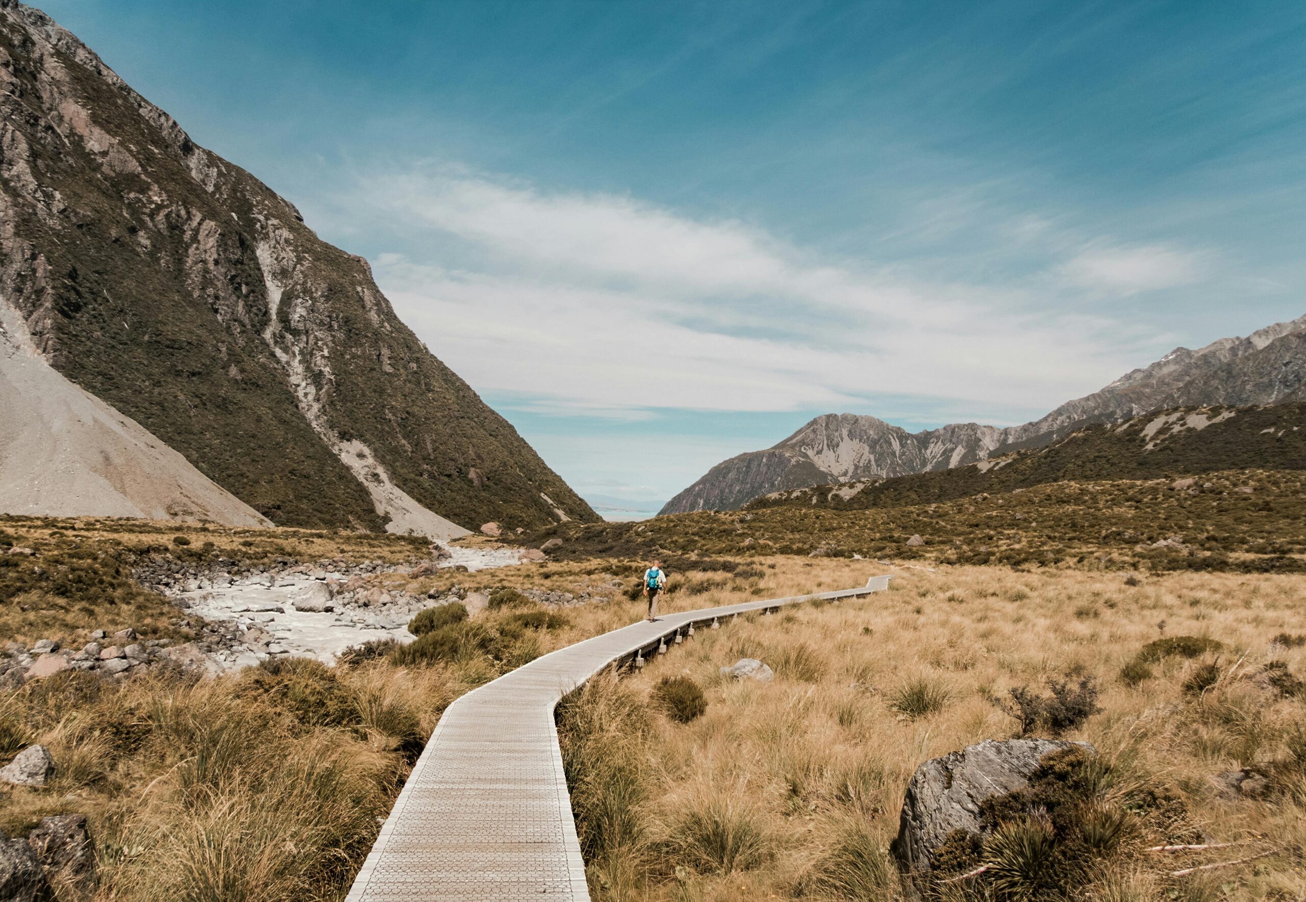 A hiker on a tranquil boardwalk path in New Zealand's mountainous landscape under a clear sky.
